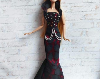 May include: A fashion doll with long blonde hair wearing a floor-length gown. The dress features a black and dark red patterned fabric, a high collar, a bodice embellished with small silver studs, and two silver chain accents at the waist.