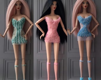 May include: Three fashion dolls with long hair and stylish dresses. The dolls have different hair colors: pink, black, and pink again. Each doll wears a beaded mini-dress in shades of green, pink, and blue, paired with matching shoes and accessories.