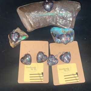 May include: Two pairs of heart-shaped earrings with iridescent glitter embedded in black resin. The earrings are displayed on brown card stock with the text "Handmade by JS".