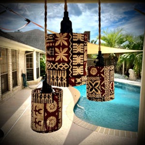 May include: Three cylindrical pendant lights hanging from braided tan cords. Each light features a dark red fabric shade with a repeating cream-colored geometric and floral pattern. The fixtures have black sockets and are suspended at varying heights.