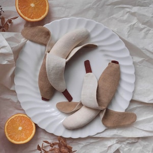 Op de afbeelding: Een stilleven met handgemaakte vilten bananen op een wit bord. De bananen zijn beige met bruine schilaccenten. Sinaasappelschijfjes en gedroogde bladeren voegen kleur en textuur toe aan de scène, tegen een achtergrond van gekreukt wit papier.