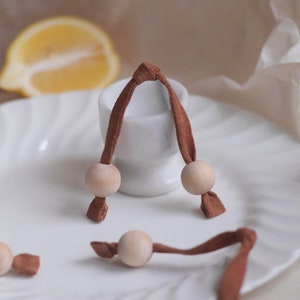 May include: A set of wooden beads strung on brown fabric cords. The beads are arranged in a circle on a white plate with a white ceramic egg cup in the centre.