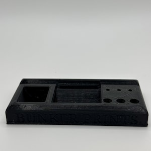 May include: Black 3D printed organizer with a small rectangular compartment, a larger rectangular compartment, and five circular compartments. The organizer has the text "BUNKER PRINTS" printed on the front.