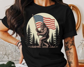 Puede incluir: Camiseta negra con un gráfico de un Bigfoot sosteniendo una bandera estadounidense en un entorno de bosque.