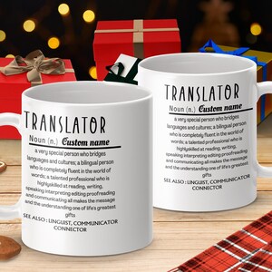 Op de afbeelding: Witte keramische koffiemok met een zwart ontwerp dat het woord "Vertaler" definieert als een heel bijzondere persoon die talen en culturen verbindt. De mok heeft een zwart handvat en staat op een rood-zwart geruit oppervlak.