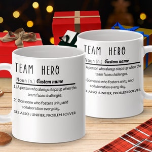 May include: White ceramic mug with a black design that reads "TEAM HERO Noun (n.) Custom name 1.) A person who always steps up when the team faces challenges. 2.) Someone who fosters unity and collaboration every day. SEE ALSO: UNIFIER, PROBLEM SOLVER"