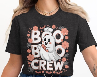 May include: Dark grey t-shirt with a Halloween graphic. The design features the words "BOO CREW" in white, with a cute ghost wearing a nurse's cap and holding a bandage. The design is surrounded by orange flowers, bats, and spiderwebs.