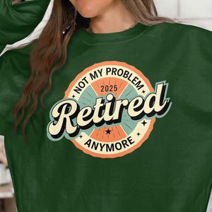Op de afbeelding: Groen sweatshirt met een retro-stijl cirkelvormige afbeelding met de tekst "Not My Problem 2025 Retired Anymore".