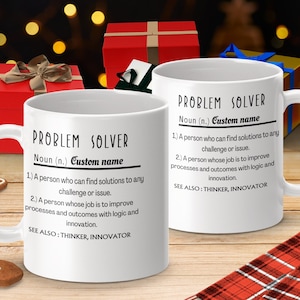 May include: Two white mugs with black text that reads "PROBLEM SOLVER" and a definition of the word. The mugs are on a wooden surface with a red and white plaid fabric and a gingerbread cookie.