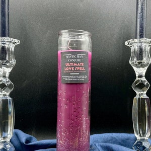 Könnte beinhalten: Eine rosa Kerze mit dem Text "Mystic Man Conjure Ultimate Love Spell" auf dem Etikett. Die Kerze steht auf einer blauen Oberfläche mit zwei schwarzen Kerzen in Kristallkerzenhaltern auf beiden Seiten.