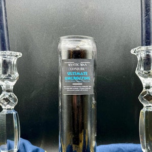 Könnte beinhalten: Eine schwarze Kerze mit einem Etikett mit der Aufschrift "Mystic Man Conjure Ultimate Uncrossing Mystic Man Supplies" in einem Glasgefäß auf einer blauen Oberfläche mit zwei Kristallkerzenhaltern und einem grünen Kristall.