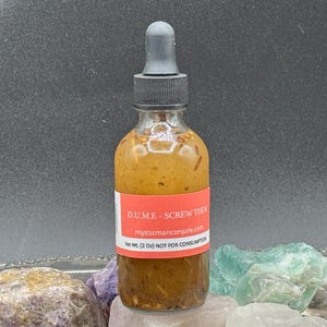 May include: A small, clear glass bottle with a brown liquid inside. The bottle has a red label with the text "D.U.M.E. - Screw Them" and the website "mysticmanconjure.com". The bottle is sitting on a surface with several colorful crystals.