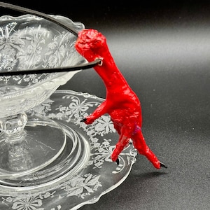 May include: A red chicken foot pendant necklace with a black cord. The pendant is hanging over a clear glass bowl with a floral design.