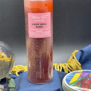 May include: A red and gold candle with the text "Mystic Man Conjure Lucky Legal Eagle MysticManConjure.etsy.com" on the label. The candle is sitting on a blue surface with a crystal ball and tarot cards.
