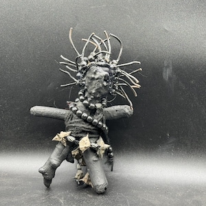 May include: A dark grey sculpture of a humanoid figure. The figure has a textured head with wire-like protrusions, a beaded necklace, and is adorned with leaf-like accents. The figure is posed on a dark background.