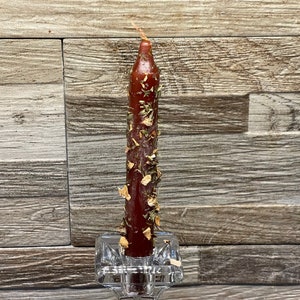 May include: A burgundy candle with dried herbs and flowers embedded in the wax, held in a clear glass holder. The background is a grey-toned wooden wall. The candle is approximately 15 cm tall.