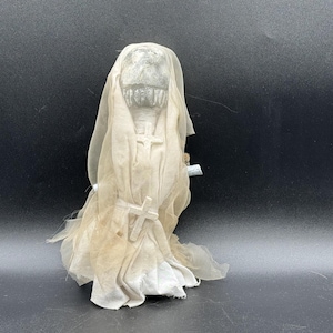 May include: A doll with a grey, textured, skull-like head featuring prominent teeth, draped in layers of sheer, off-white, distressed fabric. Two small, light-coloured cross ornaments are attached to the front of the fabric layers.