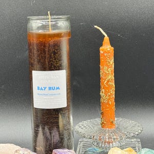May include: A large brown candle in a glass jar with a label that reads "Bay Rum" next to a smaller orange candle decorated with herbs. The candles are on a dark background with crystals.