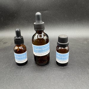 May include: Three amber glass bottles with black droppers and caps. Each bottle has a white label with blue text. The labels read "Command, Control & Obey" and "NOT FOR CONSUMPTION". The bottles are arranged on a dark surface.