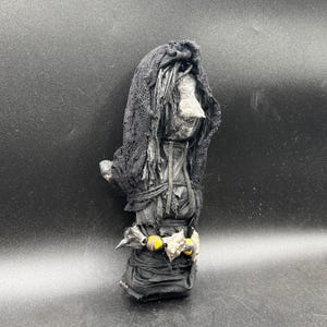 May include: A handcrafted black doll with a hooded head covering. The doll features a grey face and is wrapped in black fabric. A decorative band with yellow beads and silver accents encircles the doll's waist.