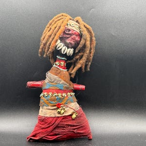 May include: A handcrafted doll with brown dreadlocks, a dark red face, and a row of white cowrie shells across the mouth. The doll wears layered fabric in red, blue, and tan, accented with gold-toned sequins, a red safety pin, and small bells.