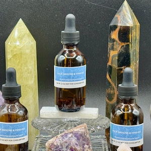 Puede incluir: Tres botellas de vidrio marrón con cuentagotas negros, cada una con una etiqueta azul que dice "FUCK AROUND & FIND OUT". Las botellas están rodeadas de cristales y piedras preciosas, incluyendo un obelisco de cuarzo amarillo y una esfera púrpura.