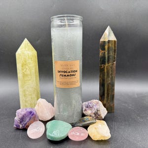 May include: An assortment of crystals and a tall, grey candle with a label reading "Mystic Man Conjure Invocation Summons." The crystals include amethyst, rose quartz, and a yellow quartz point. The candle is surrounded by various stones.