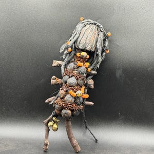May include: An abstract sculpture with a figure-like shape, crafted from natural materials. It features twigs, acorns, and seed pods, with orange bead accents. The head is made of grey fabric, and the legs are made of twigs. A unique piece of art.