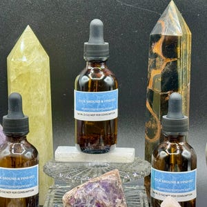 May include: Three brown glass bottles with black droppers, each with a blue label that reads "FUCK AROUND & FIND OUT". The bottles are surrounded by crystals and gemstones, including a yellow quartz obelisk and a purple sphere.