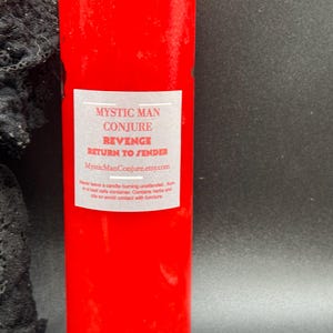 May include: A tall, cylindrical red candle with a white label featuring the text "MYSTIC MAN CONJURE REVENGE RETURN TO SENDER". The candle's label also includes a website address and safety instructions. The background is dark.