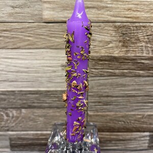 Puede incluir: Vela morada con hierbas y flores secas incrustadas en la cera. La vela está en un portavelas de vidrio transparente.