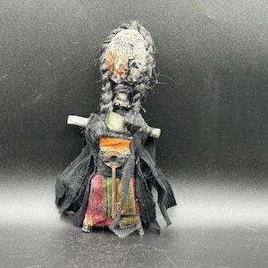 May include: A handcrafted doll featuring a textured, light-colored face with dark braided hair. The doll wears a black fabric garment over a multi-colored skirt with red, green, and yellow stripes, and a metal key is attached to the front.