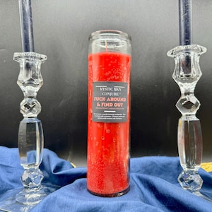 Może przedstawiać: Czerwona świeca w szklanym słoiku z czarną etykietą z napisem "Mystic Man Conjure Fuck Around & Find Out". Dwa przezroczyste szklane świeczniki z ciemnoniebieskimi świecami flankują świecę. Przedmioty znajdują się na niebieskiej tkaninie.