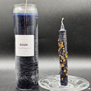 Può includere: Due candele blu scuro. Una è una candela alta e cilindrica in barattolo con un'etichetta che dice "OGUN". L'altra è una candela più piccola e decorata su un piattino di vetro. La candela più piccola ha abbellimenti color oro.