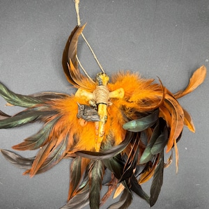 May include: A handcrafted decorative piece featuring a central cross-shaped element wrapped in twine, adorned with a small figurine. The piece is surrounded by vibrant orange and dark green feathers, creating a unique, eye-catching design.