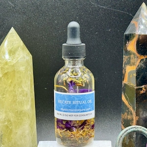 May include: A clear glass bottle with a black dropper top filled with a golden liquid and purple flowers. The bottle label reads "Hecate Ritual Oil" and "myabicmanconjure.com". The bottle is sitting on a white surface with a yellow crystal, a black and brown crystal, and a purple crystal.