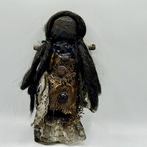 May include: A handmade doll with a dark, textured head and long black hair. The doll's clothing includes a burlap-like bodice with two buttons, layered with lace and other fabrics. The doll has a rustic, folk art aesthetic.