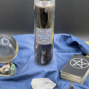 Könnte beinhalten: Eine schwarze Kerze in einem Glasgefäß mit dem Text "Mystic Man Conjure Negativity Be Gone" auf dem Etikett. Die Kerze ist umgeben von Kristallen, einer Pentagramm-Halskette und einem Tarotkartenspiel.