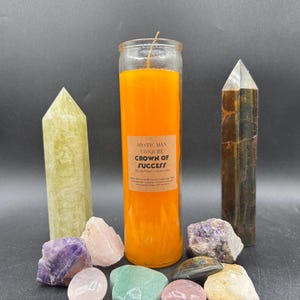 May include: An orange candle with a label reading "Crown of Success" is surrounded by crystals and crystal towers. The crystals are various colors, including purple, pink, green, and clear. The towers are green and brown.