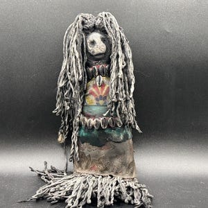 May include: A handcrafted doll featuring long, dark grey yarn hair and a face with a light-coloured, textured surface. The doll wears a layered garment with floral patterns, accented by two rows of small, white cowrie shells around the neck and waist.