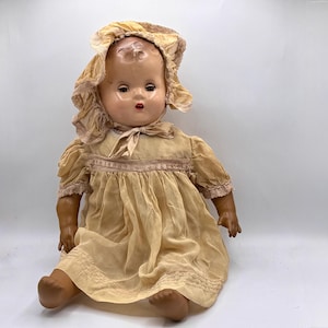 Puede incluir: Muñeca vintage con rostro pintado, vestida con un vestido amarillo claro y un gorro a juego. La muñeca tiene un rostro detallado con ojos pintados y labios rojos. El vestido presenta adornos de encaje y un lazo en el cuello. La muñeca está sentada con las piernas extendidas.
