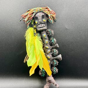 May include: A handcrafted, dark-coloured decorative figure with a skeletal face, adorned with colourful yarn hair and bright yellow feathers. The figure has a spine-like design with spiky accents.