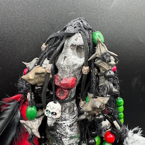 May include: A decorative figure with a textured, grey and white face, styled with black dreadlocks and various embellishments. It features a skull pendant, green, red, and black beads, and feather accents. The figure has a red mouth and is decorated with bat-like shapes.