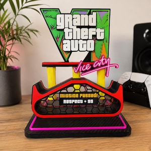 Puede incluir: Un soporte de exhibición multicapa con el logotipo de Grand Theft Auto Vice City en blanco, verde y rosa. La base incluye una estructura de panal negra con un borde rojo y un letrero que dice Mission Passed! Respect + 99 en blanco y negro.
