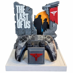 Könnte beinhalten: Ein grauer Camouflage-Videospiel-Controller ruht auf einem Ständer mit rotem Firefly-Logo. Dahinter zeigt ein steintexturierter Aufsteller den Text THE LAST OF US, die Silhouette zweier Figuren und ein rotes Banner mit weißem Firefly-Emblem.