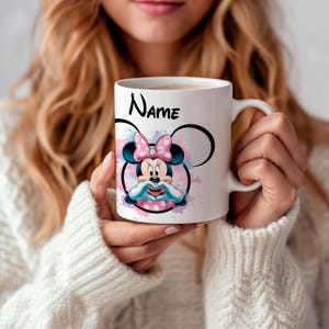 Può includere: Tazza in ceramica bianca con un design ad acquerello rosa e blu con Minnie Mouse con un fiocco rosa. La tazza ha la scritta "Name" stampata.