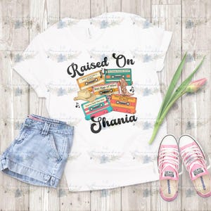 May include: White t-shirt with the text "Raised On Shania" and cassette tape graphics. The design includes a cowboy hat, cowboy boots, and musical notes. The image also shows denim shorts and pink Converse sneakers.