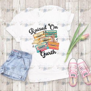 Op de afbeelding: Wit T-shirt met de tekst "Raised On Garth" en cassetteband-afbeeldingen. Het ontwerp bevat een cowboyhoed, laarzen en muzieknoten. De afbeelding toont ook een spijkerbroekshort en roze Converse sneakers.