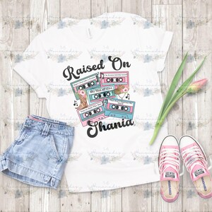 May include: White t-shirt with a graphic design featuring cassette tapes in pink and teal, floral accents, and the text "Raised On Shania." The shirt is paired with denim shorts and pink Converse sneakers.