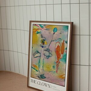 May include: A framed abstract painting titled "THE CLOWN" by Emma Stockton 2023. The painting features a colourful, whimsical style with a blue balloon, a yellow flower, and a figure resembling a clown in a blue hat.